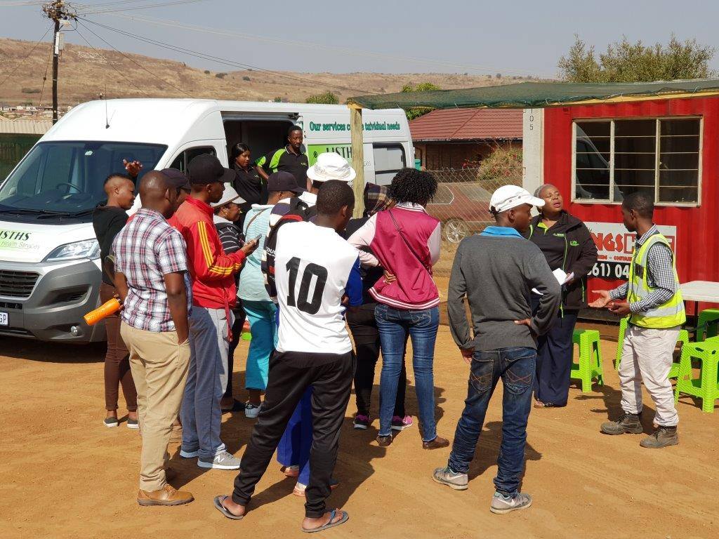 Life is True Health Services staff brief a group beside the mobile clinic at an outdoor community screening day.