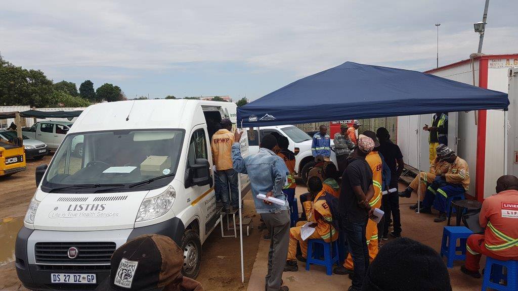 The Life is True Health Services mobile clinic is parked beside construction workers waiting with forms beneath a site canopy.
