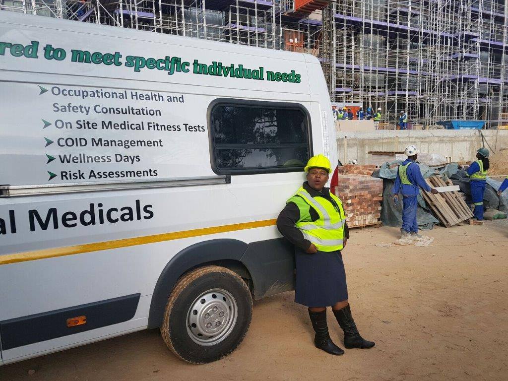 A Life is True Health Services staff member in a hard hat and high-visibility vest stands beside the mobile clinic at a construction site, with scaffolding behind her.