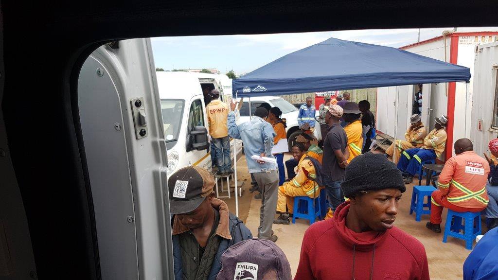 A view from inside the Life is True Health Services mobile clinic towards workers waiting with forms beneath a canopy at a workplace screening.